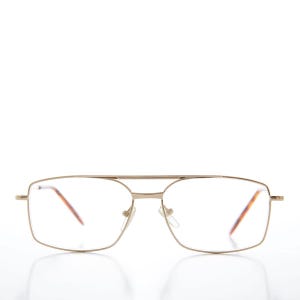 Puede incluir: Gafas de metal dorado con doble puente y lentes cuadrados. Las patillas tienen un diseño de carey. Las gafas están sobre un fondo blanco.