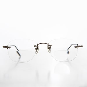 Puede incluir: Gafas de lectura sin montura con montura de metal oscuro y lentes transparentes. Las lentes tienen una sección inferior rectangular para leer. Las gafas tienen un diseño minimalista.