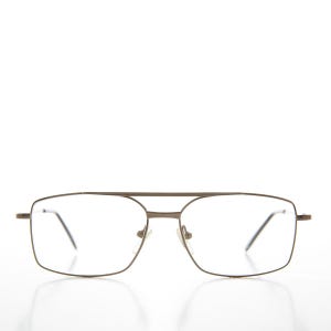 Puede incluir: Gafas con montura metálica de color bronce y forma rectangular. Las lentes son transparentes y el puente tiene un diseño de doble barra. Fondo blanco.