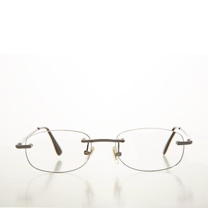 May include: Rimless spectacles with a dark grey metal frame. The lenses are oval-shaped and clear. The nose pads are a light beige colour. The temples are thin and curve slightly at the ends.