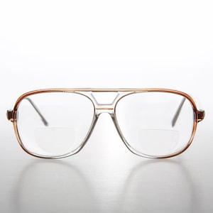 Könnte beinhalten: Transparente Aviator-Brille mit braunem Rahmen. Die Brille hat eine klare Linse mit einer kleinen Vergrößerungslinse im unteren Teil der Linse.