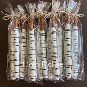 May include: Several pretzel rods dipped in white chocolate and decorated to resemble birch tree bark. Each pretzel is individually wrapped in clear cellophane and tied with twine. The text "Dela Delizie" is visible on the packaging.