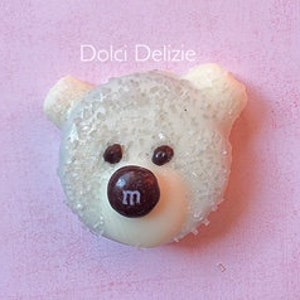 May include: A white, bear-shaped confection with a brown candy nose and two brown candy eyes. The bear's head is covered in sparkling sugar crystals. The text "Dolci Delizie" is above the bear on a pink background.
