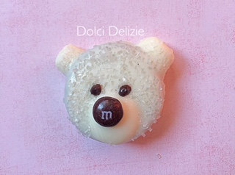 May include: A white, bear-shaped confection with a brown candy nose and two brown candy eyes. The bear's head is covered in sparkling sugar crystals. The text "Dolci Delizie" is above the bear on a pink background.