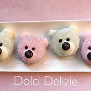 May include: Four bear-shaped treats with white, pink, and green frosting, each with a chocolate candy nose and black dot eyes, sit on a white rectangular plate. The plate rests on a pink surface. The text "Dolci Delizie" is below the plate.