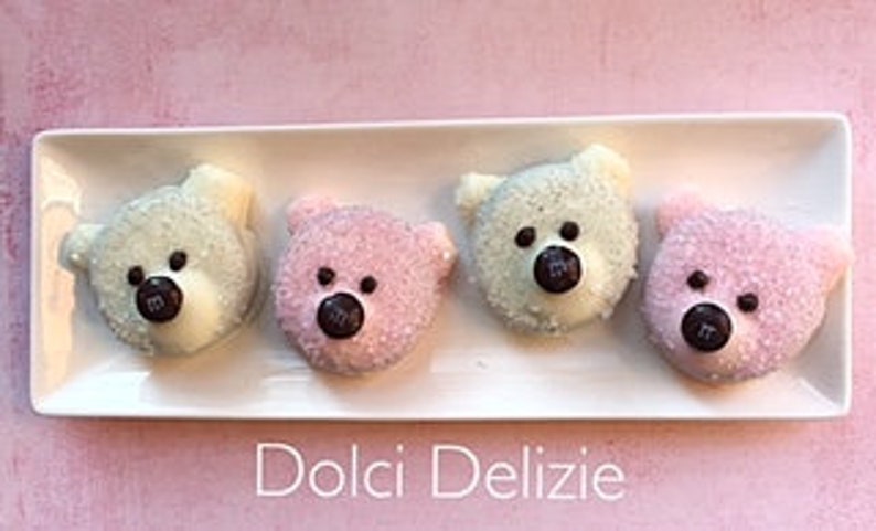 May include: Four bear-shaped treats with white, pink, and green frosting, each with a chocolate candy nose and black dot eyes, sit on a white rectangular plate. The plate rests on a pink surface. The text "Dolci Delizie" is below the plate.