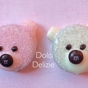 May include: Two bear-shaped candies, one pink and one white, each with a dark brown candy nose and eyes. The pink bear is covered in pink sprinkles, and the white bear is covered in white sprinkles. The candies have the letter "m" on their noses.