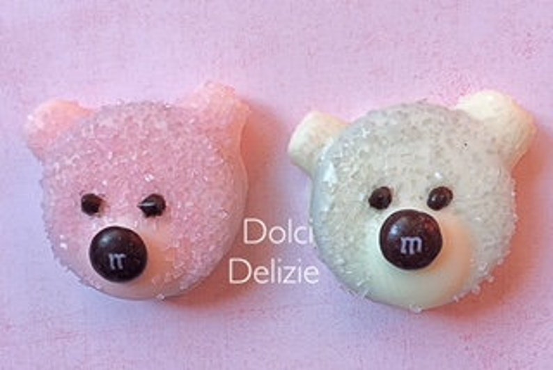 May include: Two bear-shaped candies, one pink and one white, each with a dark brown candy nose and eyes. The pink bear is covered in pink sprinkles, and the white bear is covered in white sprinkles. The candies have the letter "m" on their noses.