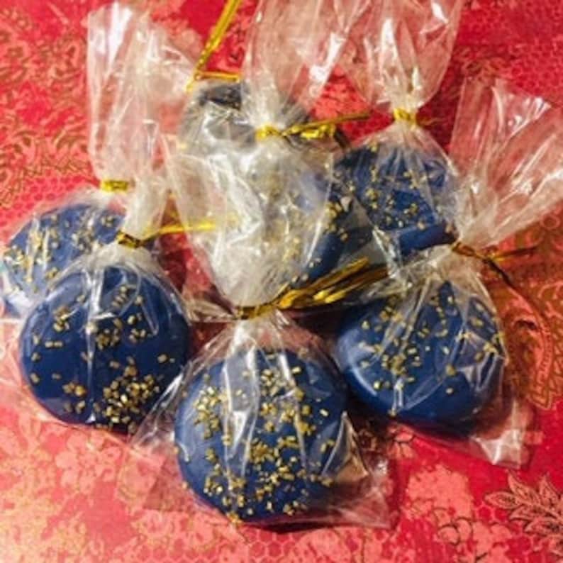 May include: Six navy blue, round, decorative ornaments with gold glitter, wrapped in clear cellophane and tied with gold string. The ornaments are arranged on a red patterned surface.