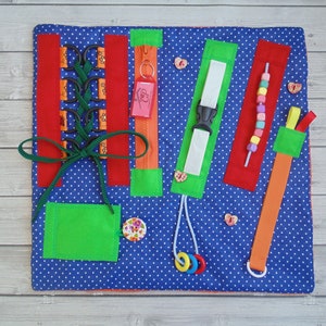 Puede incluir: Una alfombra de actividades de tela azul con varias texturas y actividades para niños. Cuenta con una cremallera, una hebilla, una actividad de cordones, una cadena de cuentas y una correa de velcro.