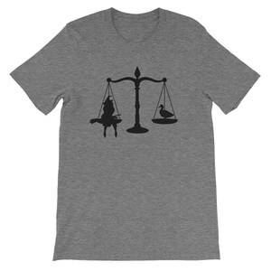 May include: A grey t-shirt featuring a black silhouette of a scale with a witch on one side and a duck on the other.