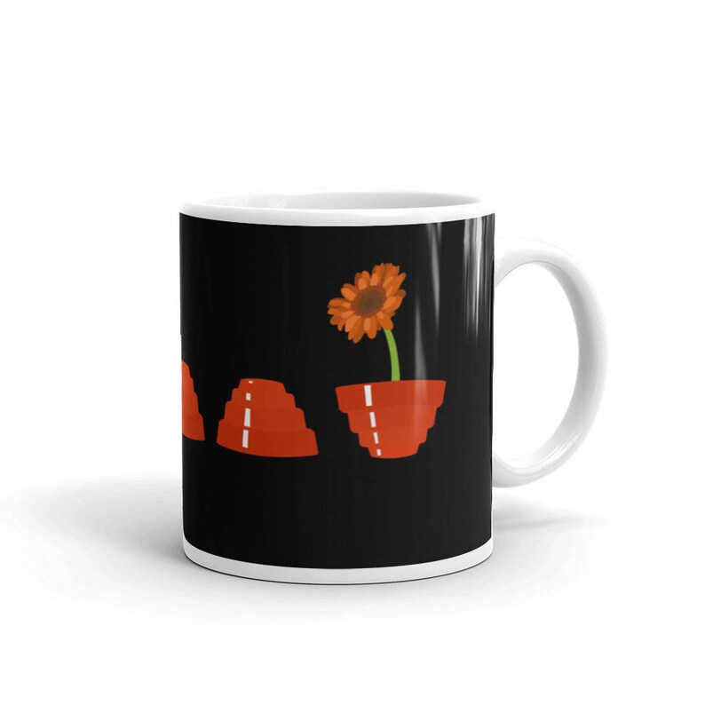 May include: A white ceramic mug with a black background featuring a stylised orange flower in a red pot. The flower is growing out of the pot and has a green stem. There are three red stacked pots to the left of the flower.