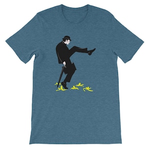 May include: Teal short-sleeved t-shirt with a black and white illustration of a man in a bowler hat, coat, and cane, slipping on yellow banana skins. The design is a humorous take on a classic slapstick comedy scene.