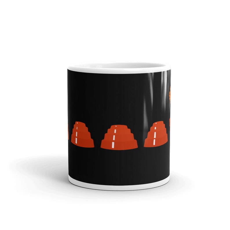 May include: A white ceramic mug with a black background and a design of three red stacked cones with white lines on a black background.