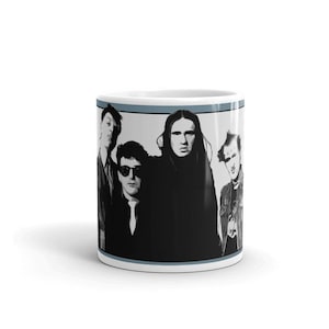 Può includere: Una tazza in ceramica bianca con un'immagine in bianco e nero di una band. I membri della band indossano tutti abiti scuri e hanno i capelli acconciati in stile punk rock. La tazza ha una striscia blu intorno alla parte superiore.
