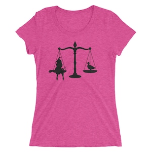 May include: A pink women's t-shirt featuring a black silhouette of a scale with a witch on one side and a duck on the other.