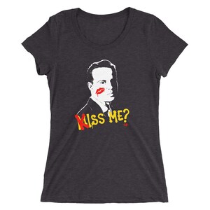 Può includere: Una maglietta grigio scuro con un'illustrazione in bianco e nero di un uomo con un segno di rossetto rosso sulla guancia. Il testo "Kiss Me?" è stampato in lettere gialle sotto l'illustrazione.