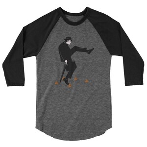 Peut inclure: Un t-shirt de baseball noir et gris avec une illustration en noir et blanc d'un homme portant un chapeau marchant sur un chemin de crottes brunes. Le t-shirt a une manche raglan noire.