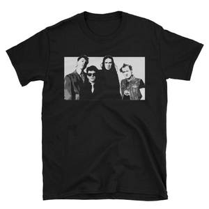 Può includere: Foto in bianco e nero di una band con quattro membri. I membri della band indossano abiti scuri e hanno i capelli corti. La foto è stampata su una maglietta nera.