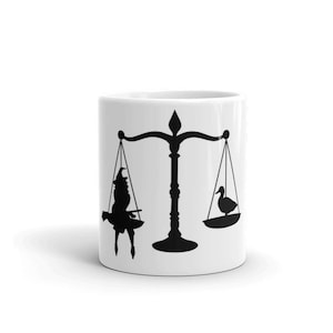 May include: White ceramic mug featuring a black silhouette of scales with a witch on one side and a duck on the other.