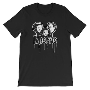Op de afbeelding: Zwart T-shirt met een witte afbeelding van drie mannengezichten en het woord "Misfits" dat druppelt in witte verf.