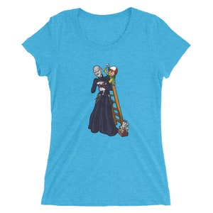 Puede incluir: Una camiseta de mujer azul turquesa con un estampado gráfico de una figura alta y oscura sosteniendo una figura pequeña en una escalera. La figura alta lleva una túnica negra y la figura pequeña lleva un atuendo verde y amarillo. El gráfico recuerda a una película de terror.