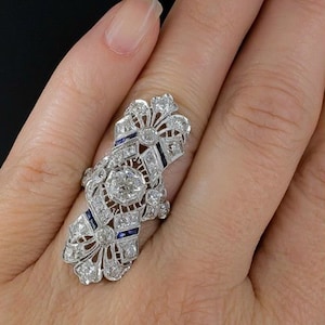 May include: A silver ring with a large, round, clear center stone surrounded by smaller clear stones. The ring has a delicate, intricate design with blue sapphire accents.