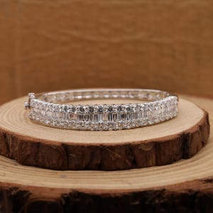 Könnte beinhalten: Ein silberfarbenes, aufklappbares Armreif-Armband mit zwei Reihen funkelnder, klarer Edelsteine. Das Armband liegt auf einer Holzoberfläche, die sein elegantes Design und seine komplizierten Details hervorhebt. Die Edelsteine sind rechteckig und rund geschliffen.