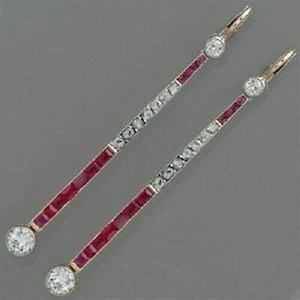 May include: A pair of gold-toned drop earrings. Each earring features a line of small, rectangular red stones, interspersed with clear, round-cut stones. A large, round, clear stone is at the top and bottom of each earring. The earrings are set against a gray background.