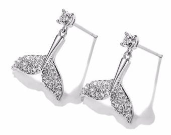 May include: A pair of silver-toned stud earrings. Each earring features a sparkling, crystal-covered mermaid tail design hanging below a small, round, clear crystal. The earrings are set against a white background.