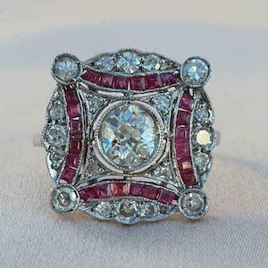 May include: An antique-style ring with a large, round, clear center stone. The ring features a square design with smaller clear stones and red stones. The ring is set in a silver-colored metal.