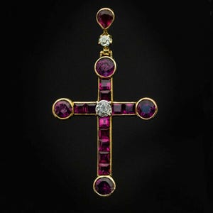May include: A gold-toned cross pendant featuring a central diamond and numerous deep red gemstones. The cross has a vertical and horizontal bar design, with a teardrop-shaped gemstone at the top. The gemstones are set in a gold frame.