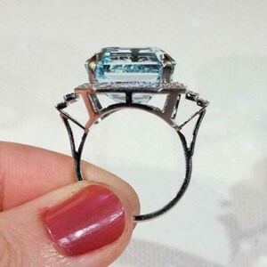 May include: A silver ring with a large, light blue, emerald-cut aquamarine gemstone surrounded by a halo of smaller, clear gemstones.
