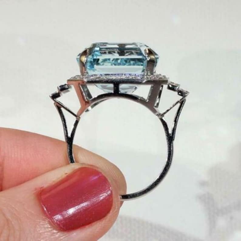May include: A silver ring with a large, light blue, emerald-cut aquamarine gemstone surrounded by a halo of smaller, clear gemstones.