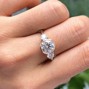 May include: A gold ring featuring three round, clear gemstones. The largest gemstone is centered, with a smaller one on each side. The ring is set in a simple, elegant design, showcasing the brilliance of the stones.