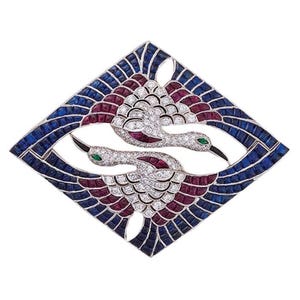 May include: A diamond-encrusted brooch featuring two stylized birds in flight. The birds have ruby and diamond bodies, emerald eyes, and black beaks. The brooch is framed by a geometric design with blue sapphires and rubies, creating a striking contrast.
