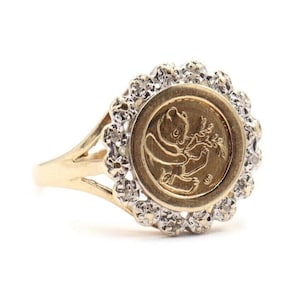 May include: A gold ring featuring a circular gold coin with a panda bear design, surrounded by a halo of small, clear gemstones. The ring has a split shank design.