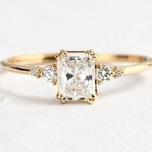 Könnte beinhalten: Ein goldener Verlobungsring mit einem großen, rechteckigen, klaren Diamanten und zwei kleineren Diamanten auf jeder Seite.