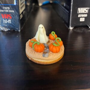 May include: A Halloween-themed figurine featuring a white ghost figure surrounded by orange pumpkins with green stems. The pumpkins are set on a circular base with a straw-like texture. The figurine is a miniature decorative item.