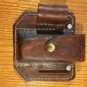 May include: A brown leather holster with a stitched edge and a brass button. The holster has a loop for a belt and a separate compartment for a small item.