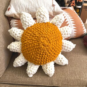 May include: A crocheted sunflower pillow with a yellow center and white petals. The pillow is sitting on a brown couch.