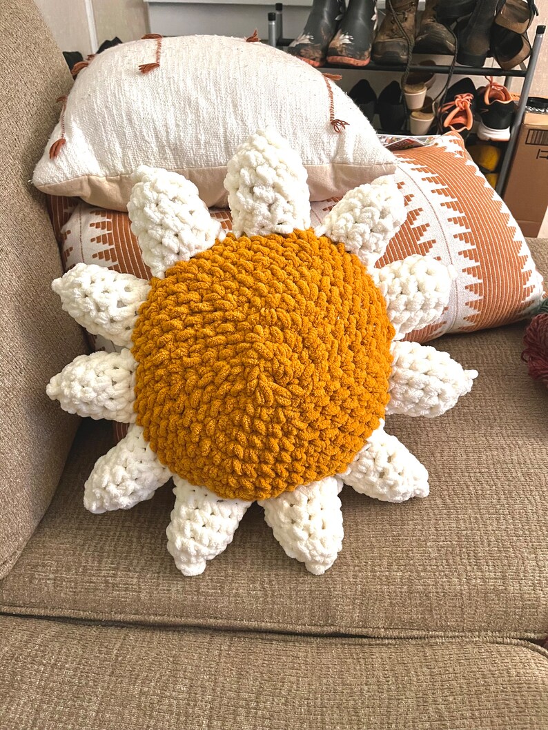 May include: A crocheted sunflower pillow with a yellow center and white petals. The pillow is sitting on a brown couch.