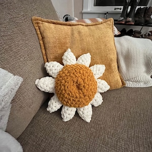 May include: A crocheted sunflower pillow with a white center and yellow petals. The pillow is sitting on a brown couch.