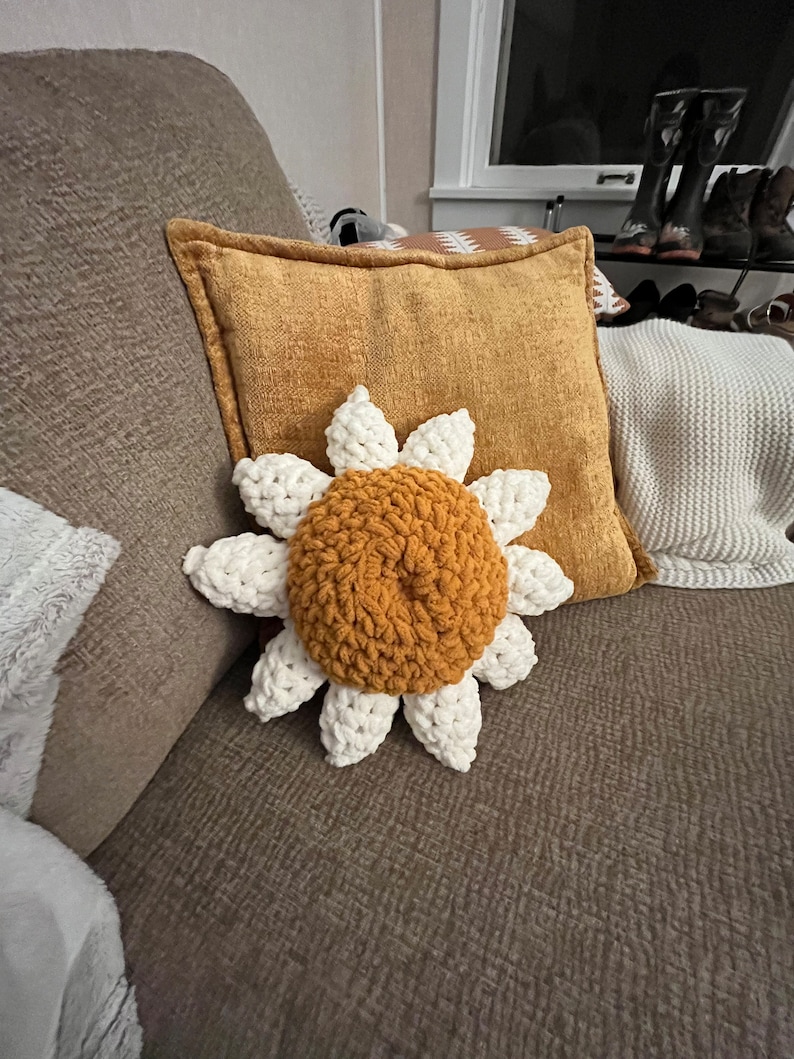 May include: A crocheted sunflower pillow with a white center and yellow petals. The pillow is sitting on a brown couch.