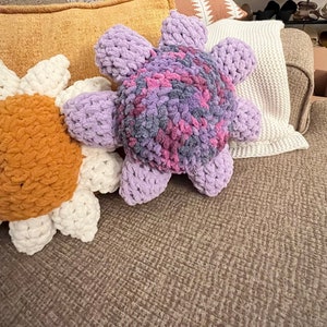 May include: Two crocheted flower-shaped pillows, one in a light purple and pink color with a gray center, and the other in a white and orange color. The pillows are on a brown couch.