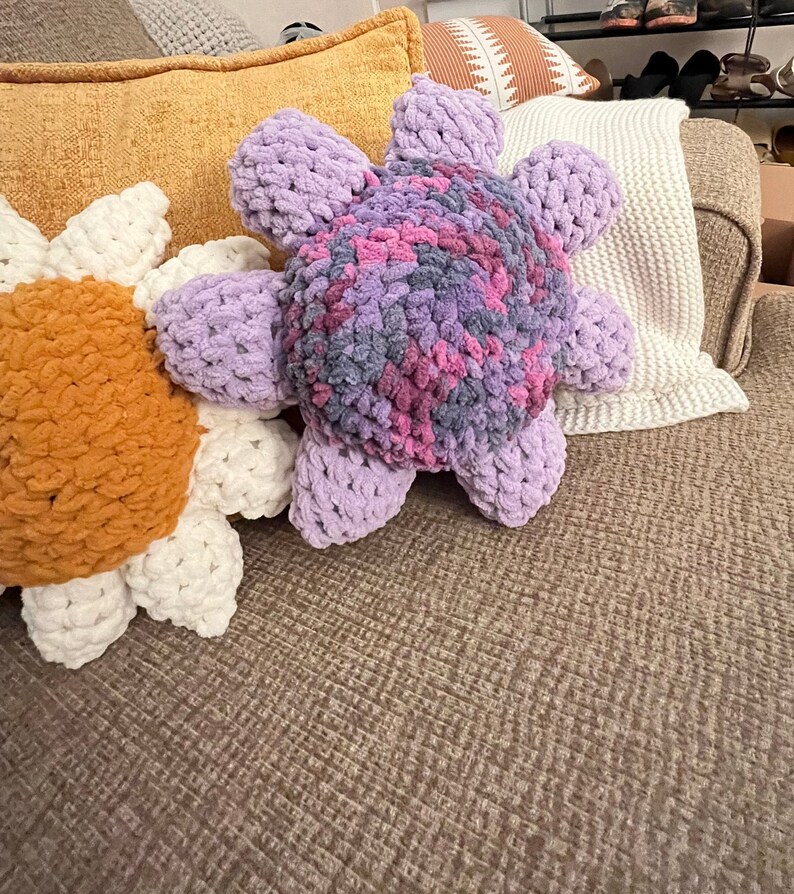 May include: Two crocheted flower-shaped pillows, one in a light purple and pink color with a gray center, and the other in a white and orange color. The pillows are on a brown couch.
