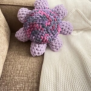 May include: A plush purple and pink octopus-shaped pillow with a textured surface. The pillow is lying on a white knitted blanket.