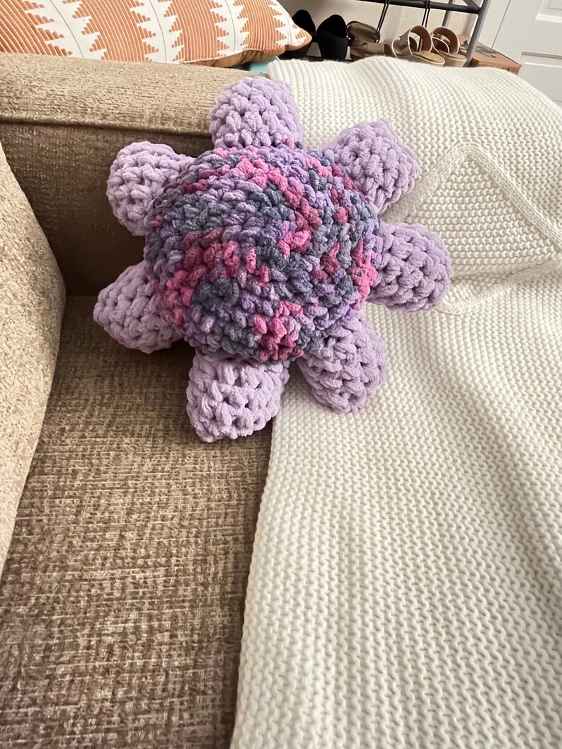 May include: A plush purple and pink octopus-shaped pillow with a textured surface. The pillow is lying on a white knitted blanket.