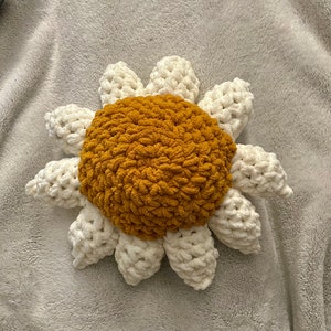 May include: A crocheted sunflower pillow with a white center and yellow petals. The pillow is lying on a white fuzzy blanket.