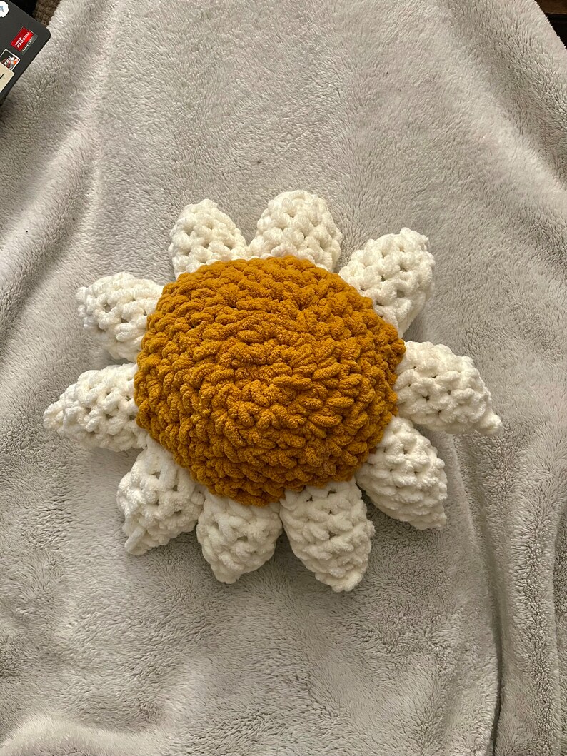 May include: A crocheted sunflower pillow with a white center and yellow petals. The pillow is lying on a white fuzzy blanket.